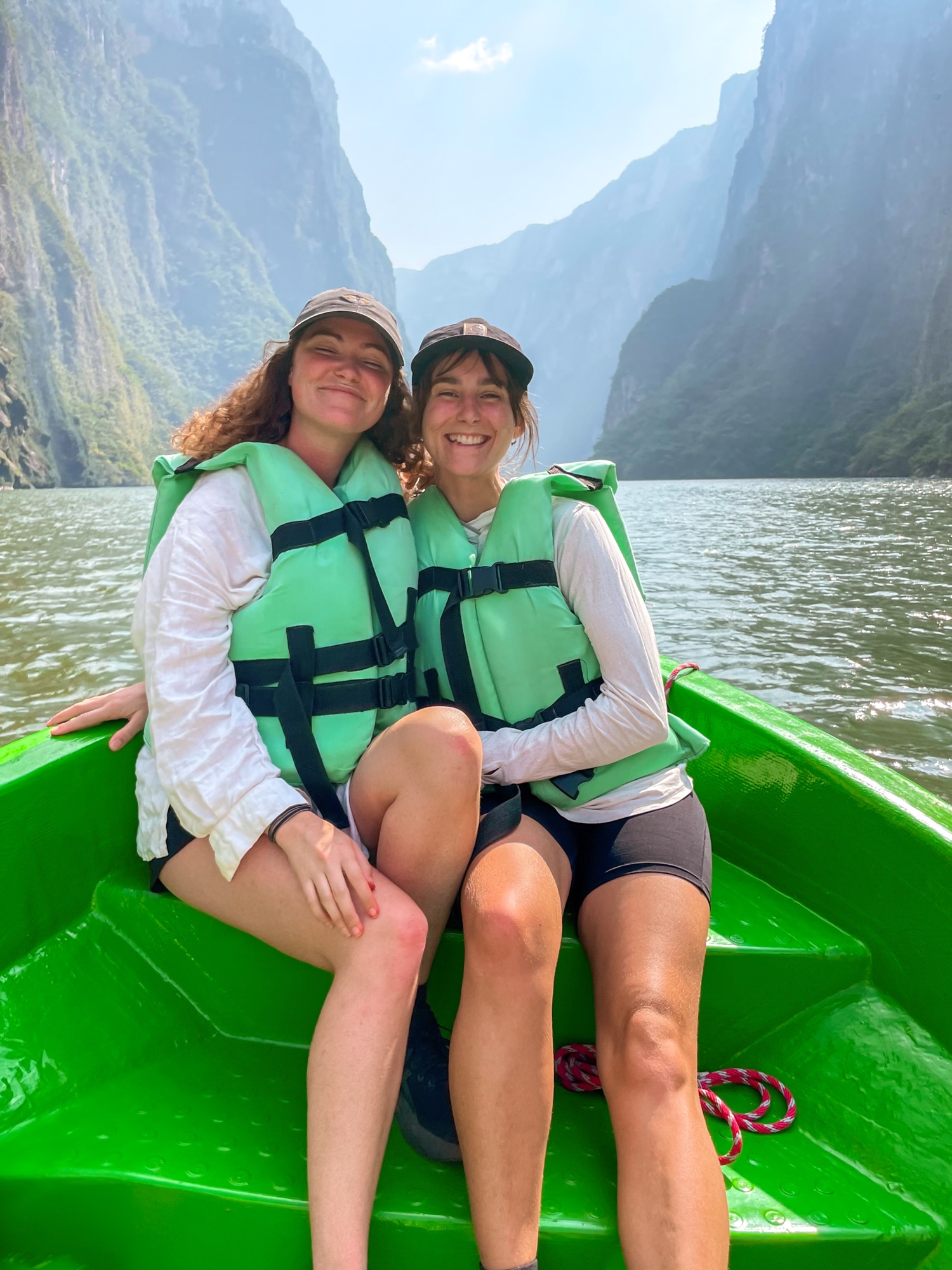 Two girls pose for a photo on a green boat with the river and steep walls of the canyon behind them
