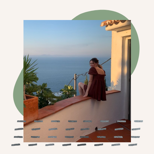 Ren sitting on a balcony, her face is turned to the camera but her body faces the sea and sky