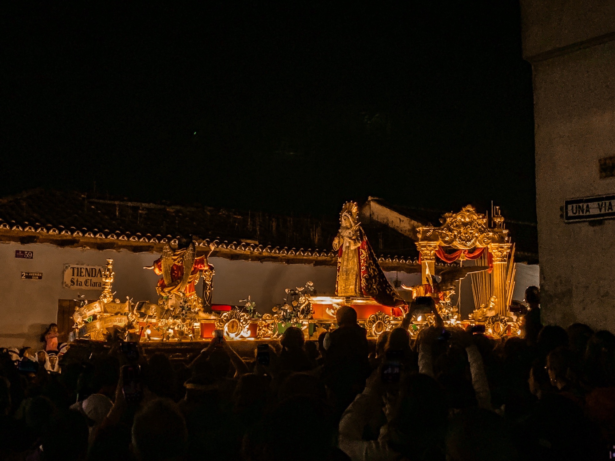 At night, a crowd (in shadows) looks onto a procession for Semana Santa with the float brightly lit and baring religious imagery