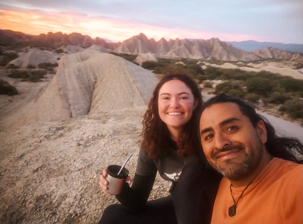 my friend and I sitting on a hill, drinking mate, smiling for the photo with the sunset and rock formations behind us