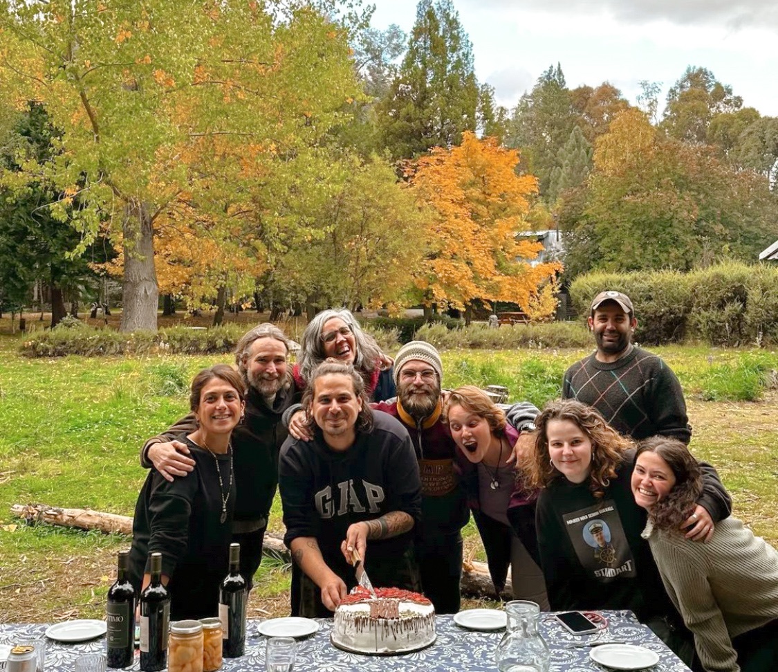 a group of volunteers pose for a photo while the man in the centre cuts a birthday cake. behind them is the beginning of an argentina winter with the trees turning golden