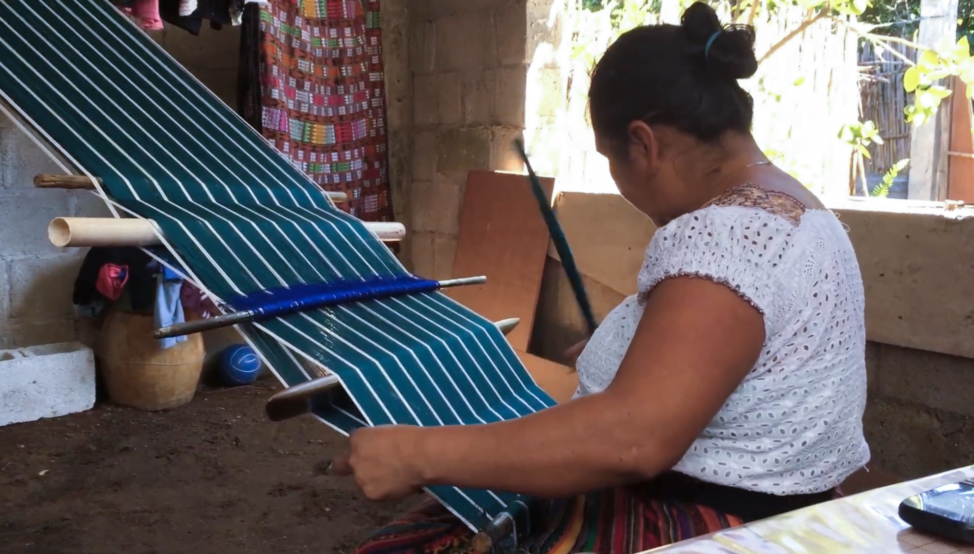 A Mayan woman wearing a white blouse faces away from the camera while using a backstrap loom to weaving Maya textiles. The design she's making is a teal-blue and white. It would take her a week to get to the end of this creation