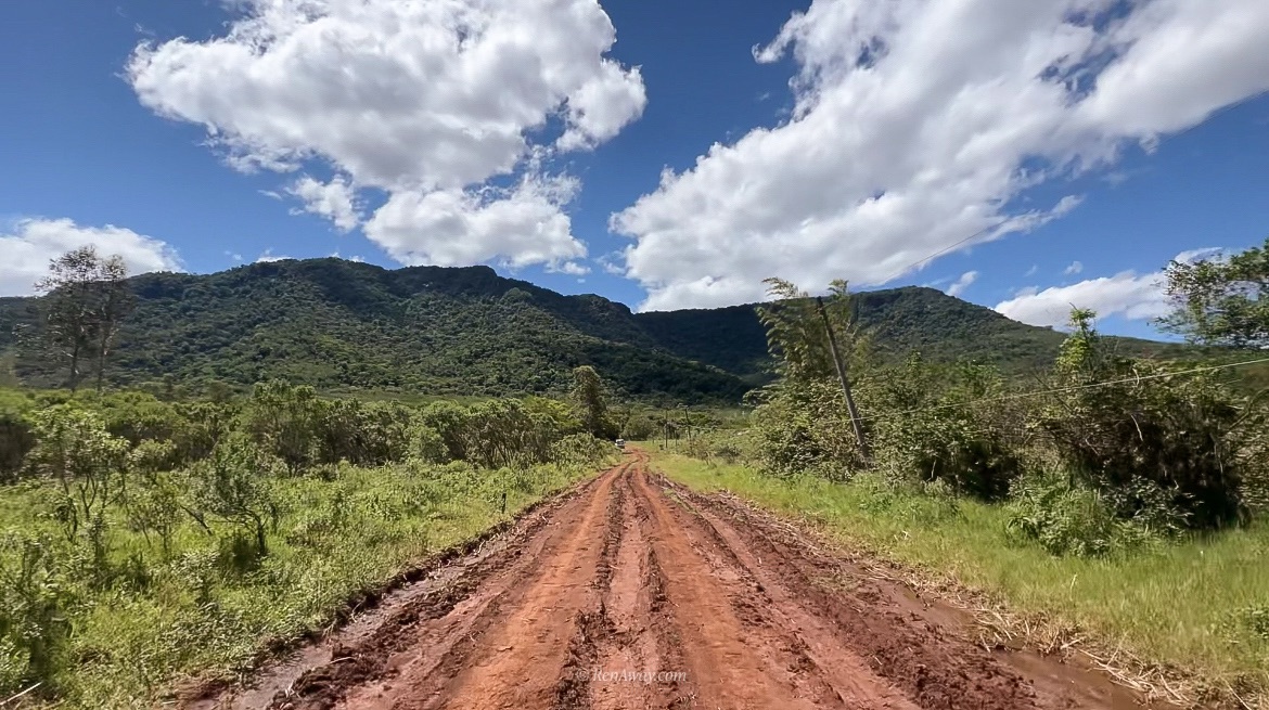 A red dirt road leading towards a small mountain range, the grass is light green and the hill is dark green from the vegetation. Its a sunny day with a few clouds